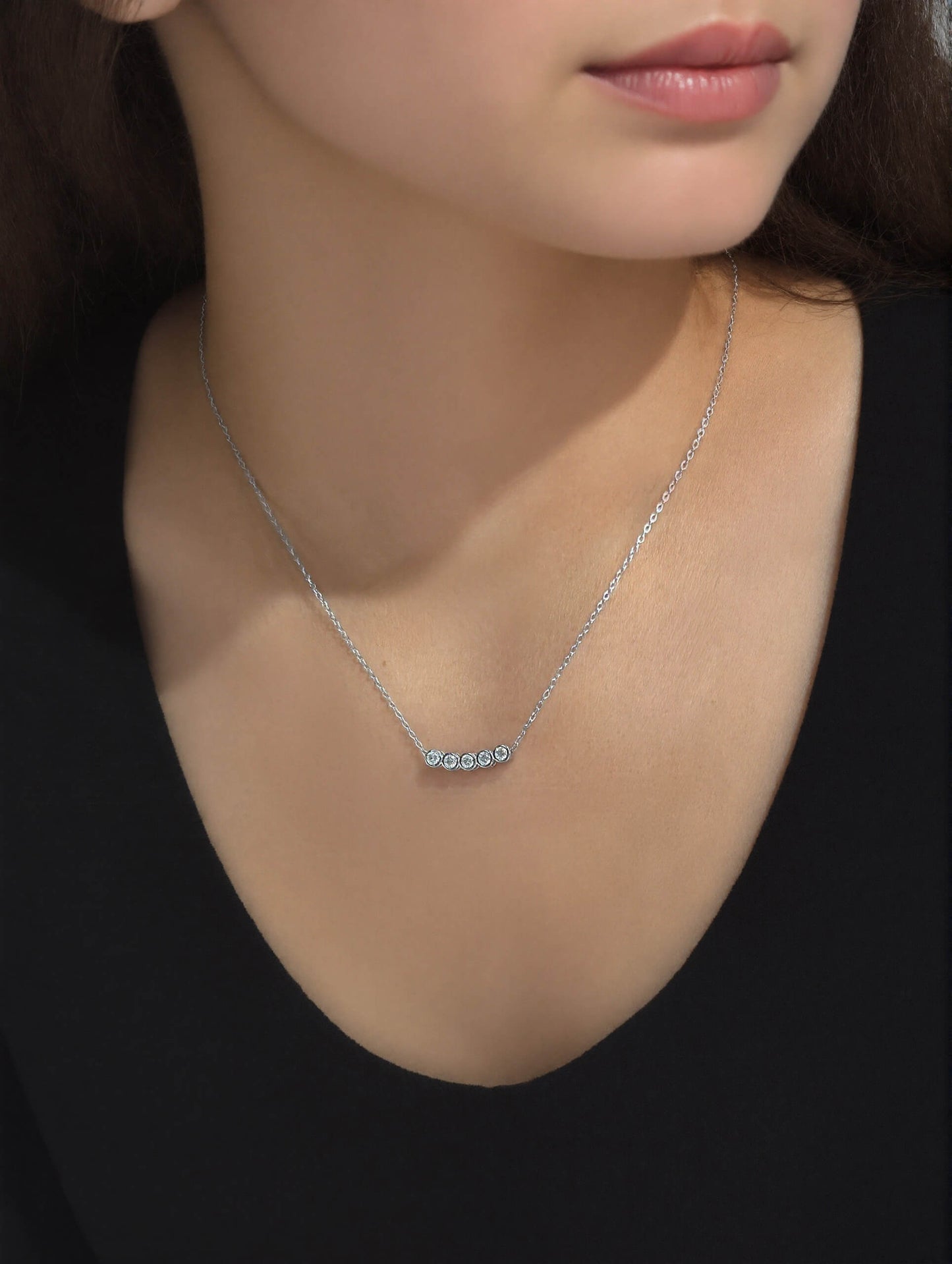 Model wearing the chic 925 Silver Dawn Necklace with a fine chain and horizontal bar pendant featuring five round bezel-set sparkling stones, styled over a cream satin top.