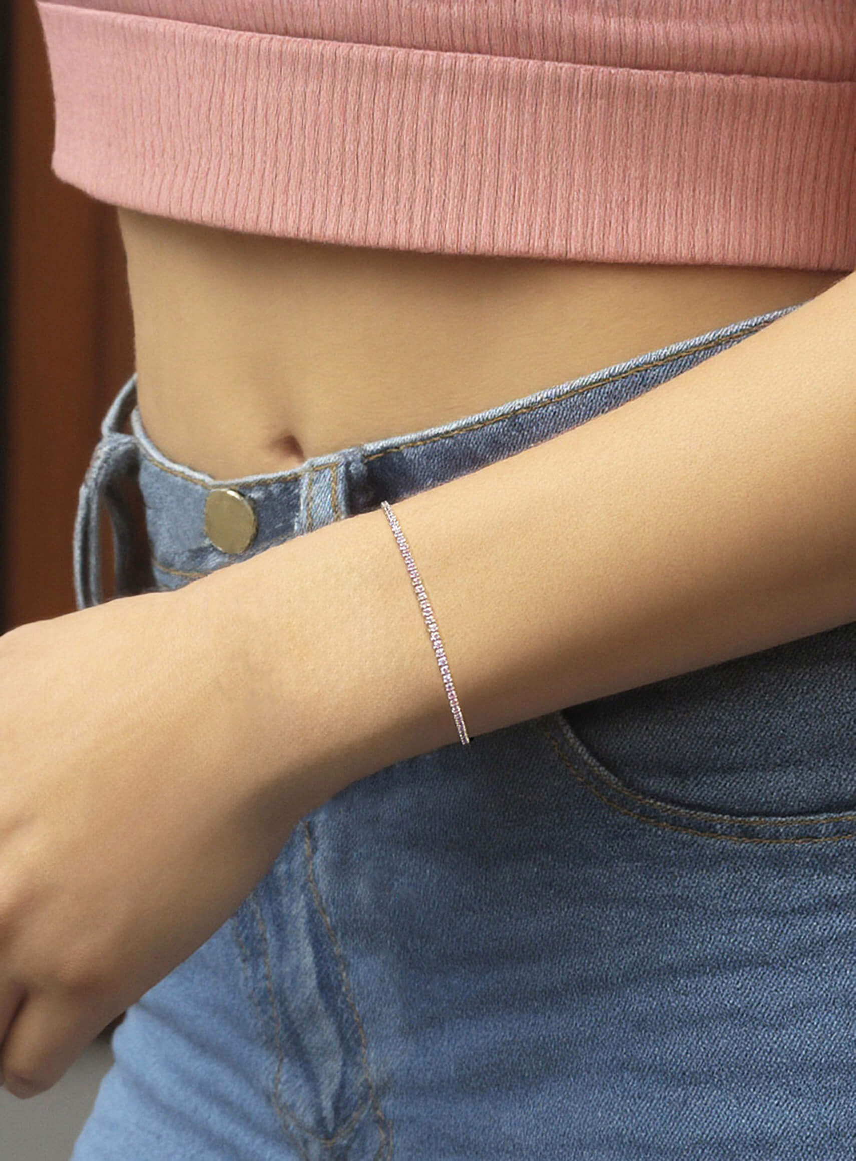 Model’s arm styled with the dainty 925 silver Amore tennis bracelet, featuring a continuous row of sparkling pink stones, paired with a pink top and blue jeans for a playful everyday look.