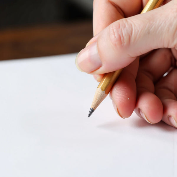 A hand holding a pencil over a blank page, representing the beginning of our brand's story and our mission to create timeless, quality jewellery.