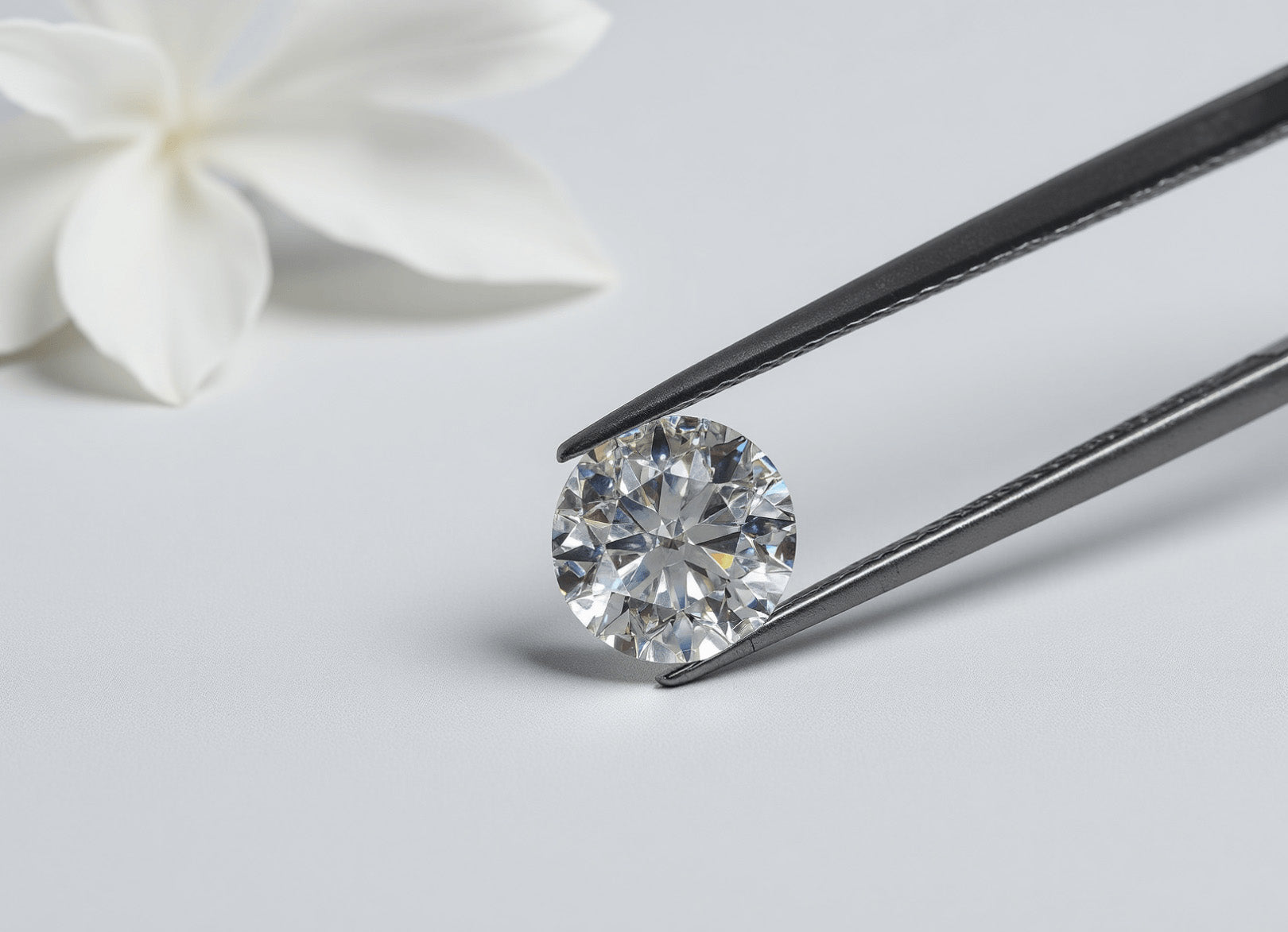 Loose premium round brilliant-cut moissanite gemstone held in jeweler's tweezers, sparkling against a white background with a soft-focus flower. 