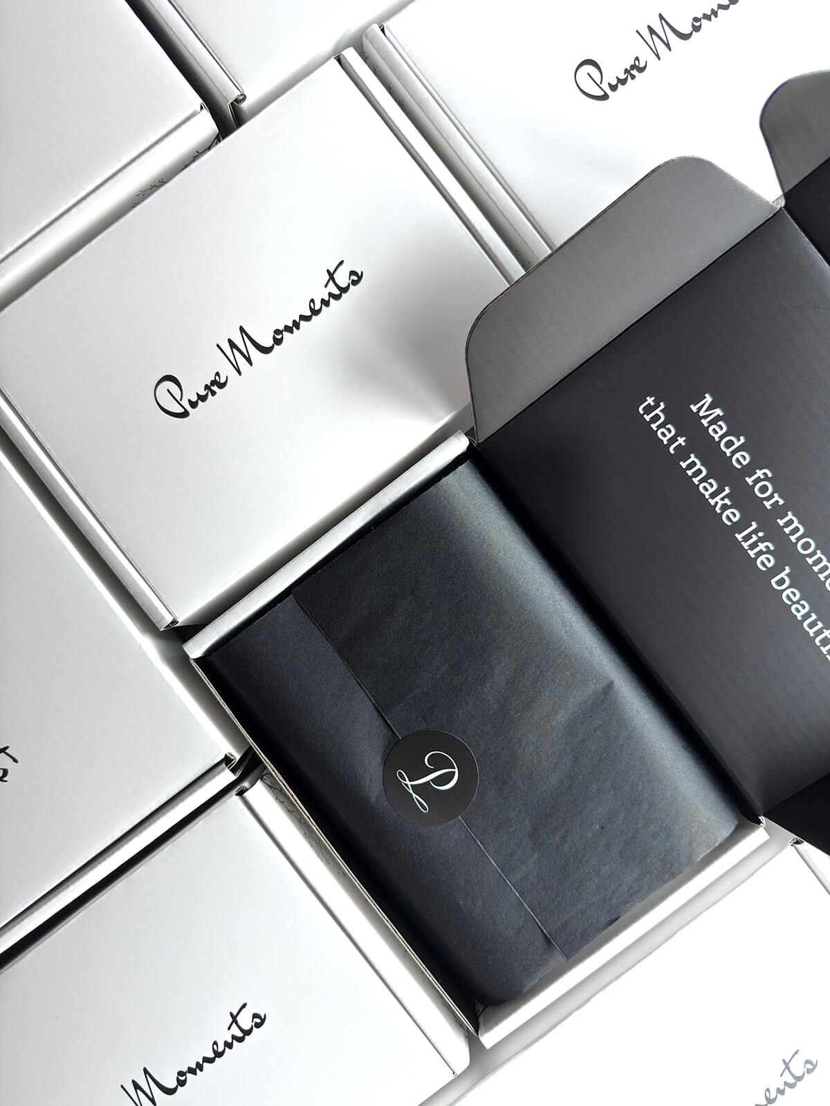 An open Pure Moments shipping box revealing the brand’s tagline, “Made for moments that make life beautiful,” printed inside the lid. Below, black tissue paper sealed with a logo sticker represents our care and commitment to satisfaction.